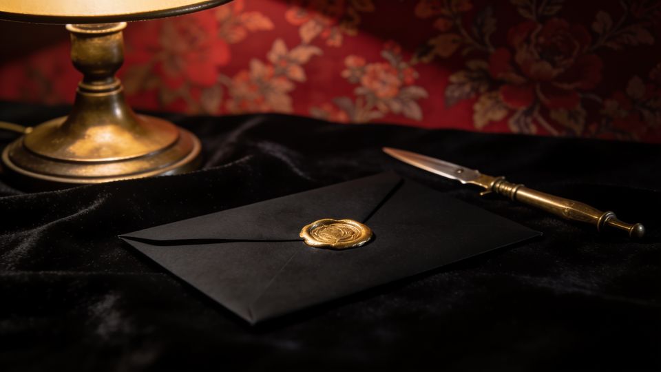 A black envelope sealed with gold wax beside a brass letter opener